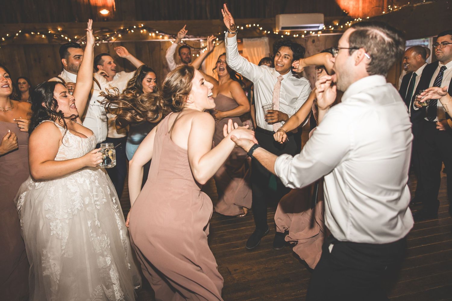 Energetic wedding reception dance floor with guests dancing under glowing string lights.