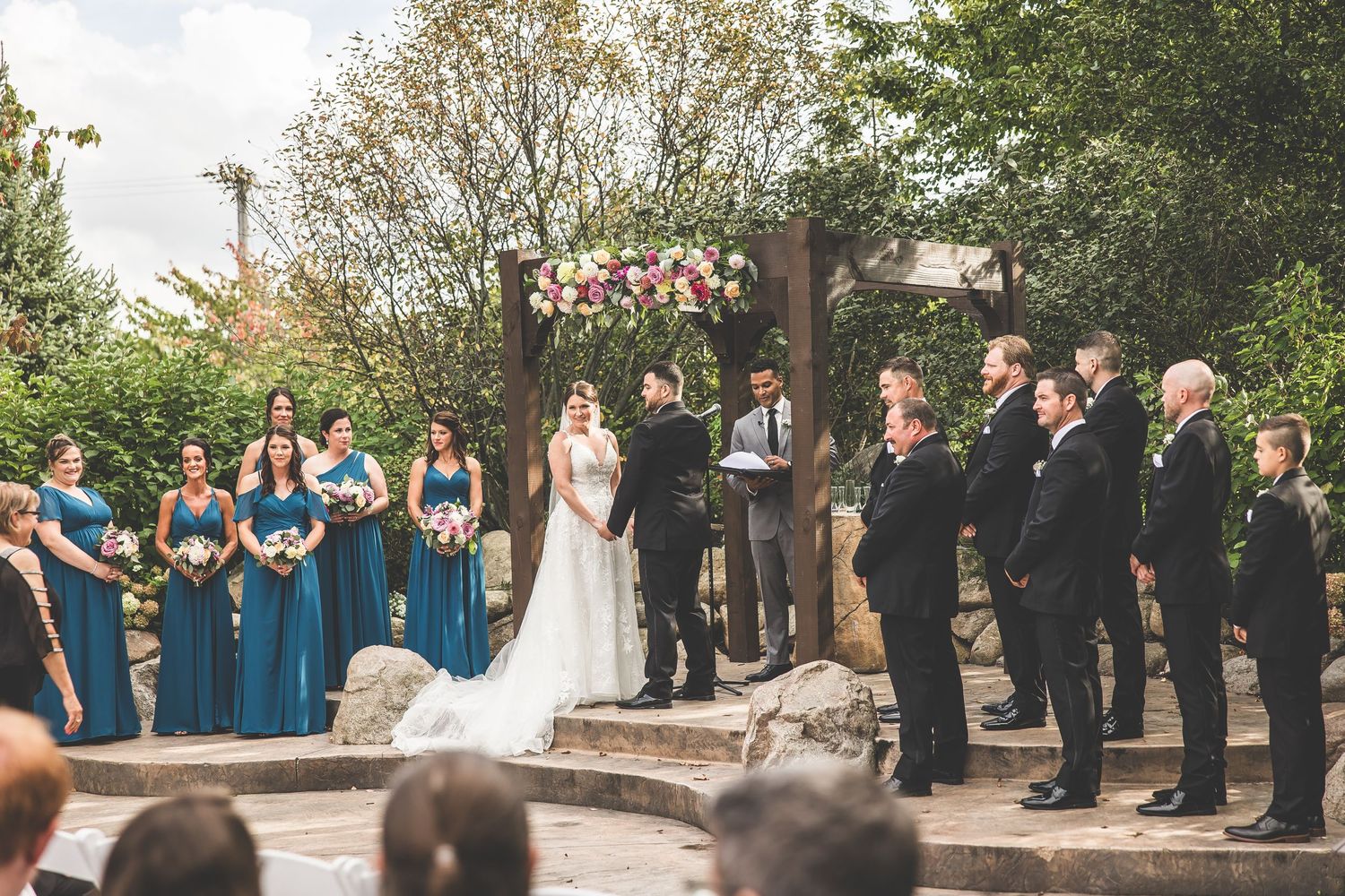 Wedding party and guests gathered outdoors during a ceremony with bridesmaids in blue dresses.