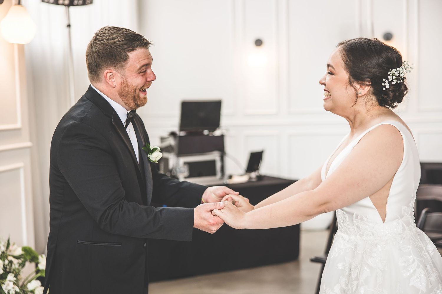 Emotional wedding first look moment captured in a bright white room with black folding chairs.