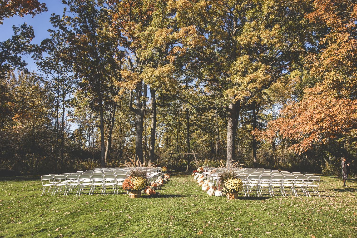 Outdoor fall wedding ceremony setup with white chairs and pumpkin decorations in a wooded setting.