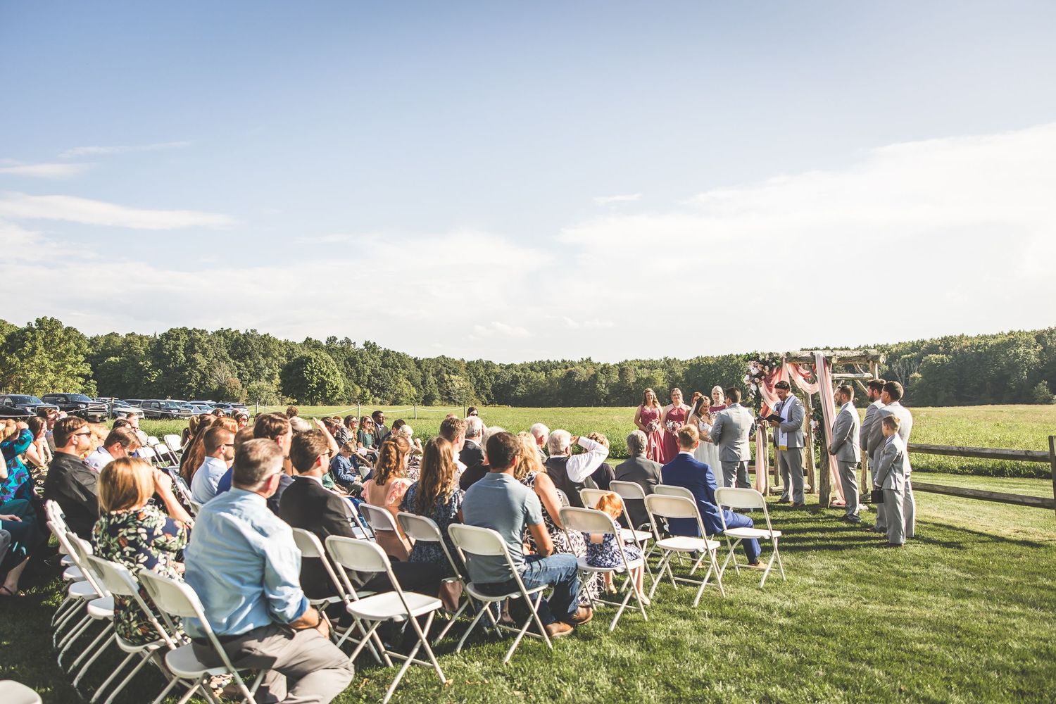 Wide view of outdoor wedding ceremony with guests seated in white chairs on grassy field.