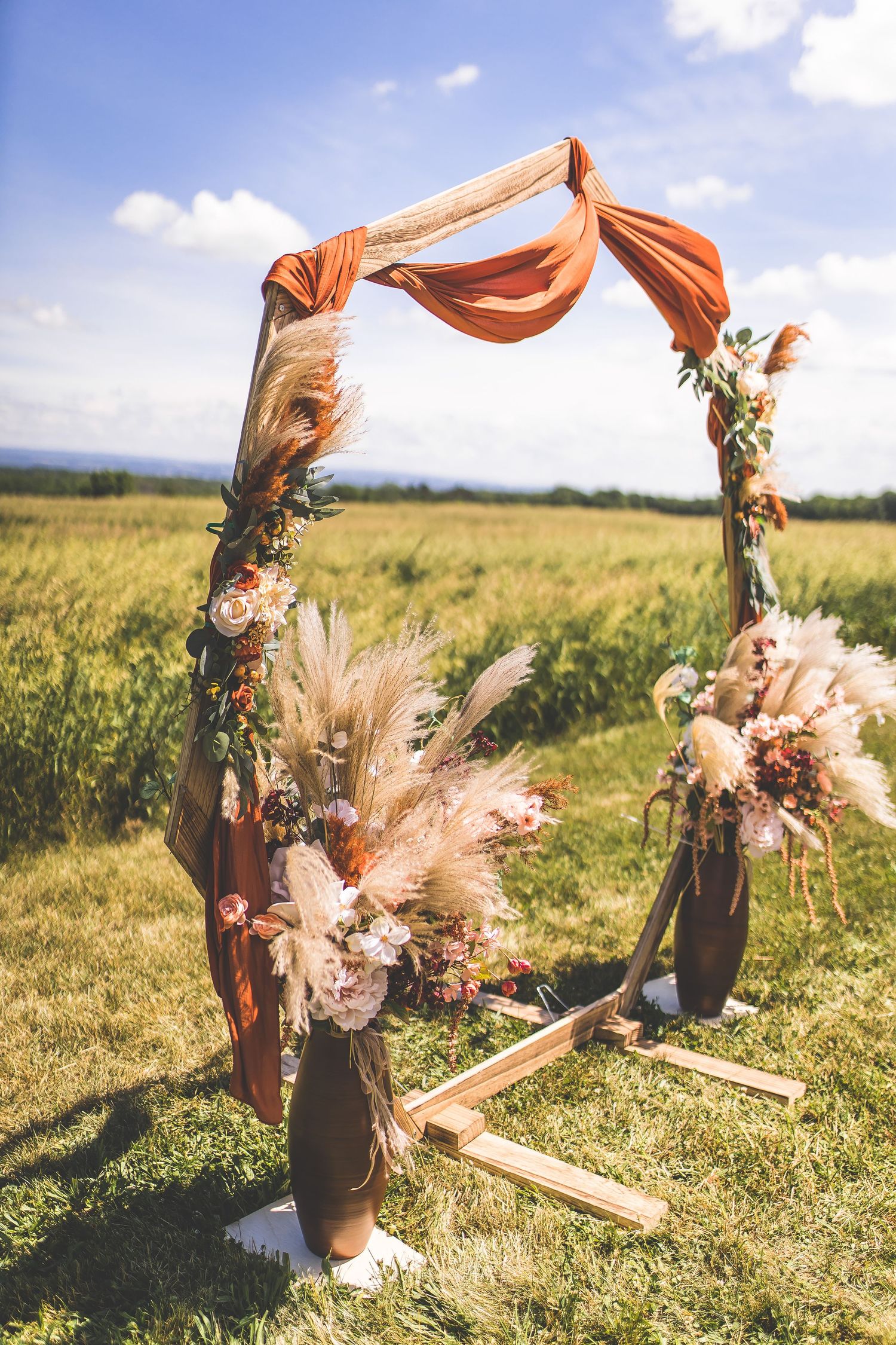 Coral fabric arch with pampas grass and floral decorations stands in a sunny meadow.