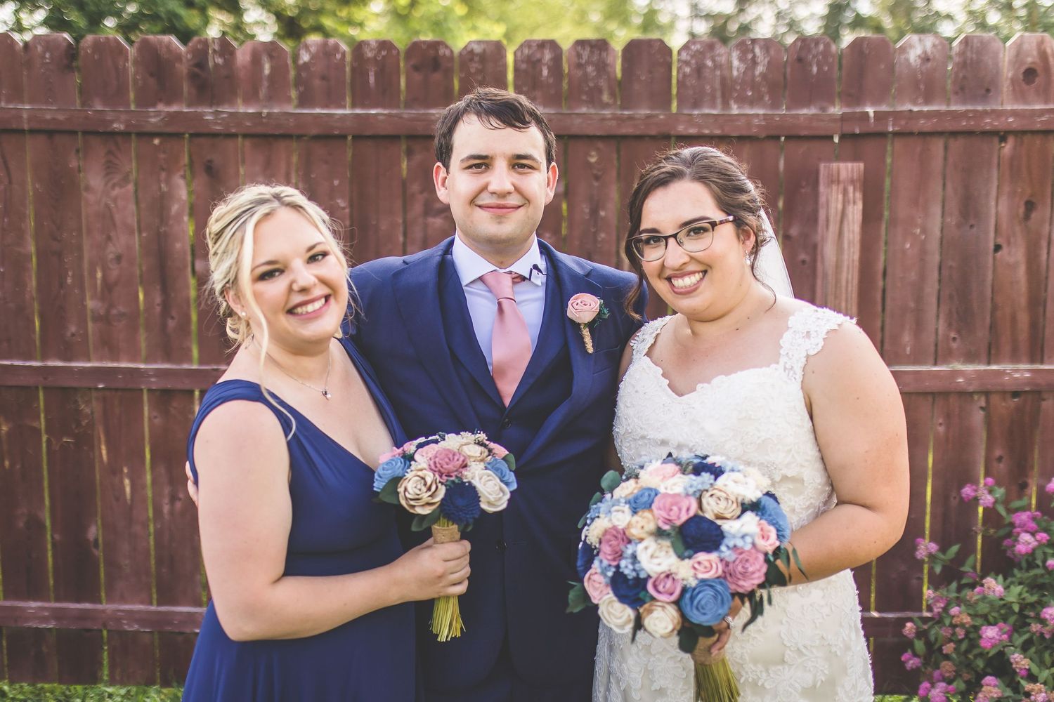 Wedding party members pose together holding blue and pink floral bouquets against a wooden fence.