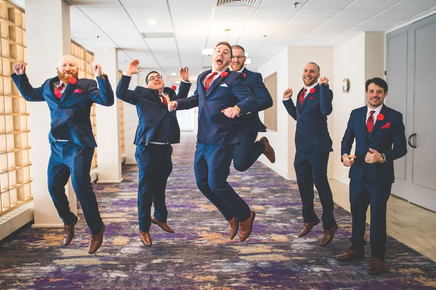 Groomsmen jumping and celebrating in navy suits inside modern hallway.