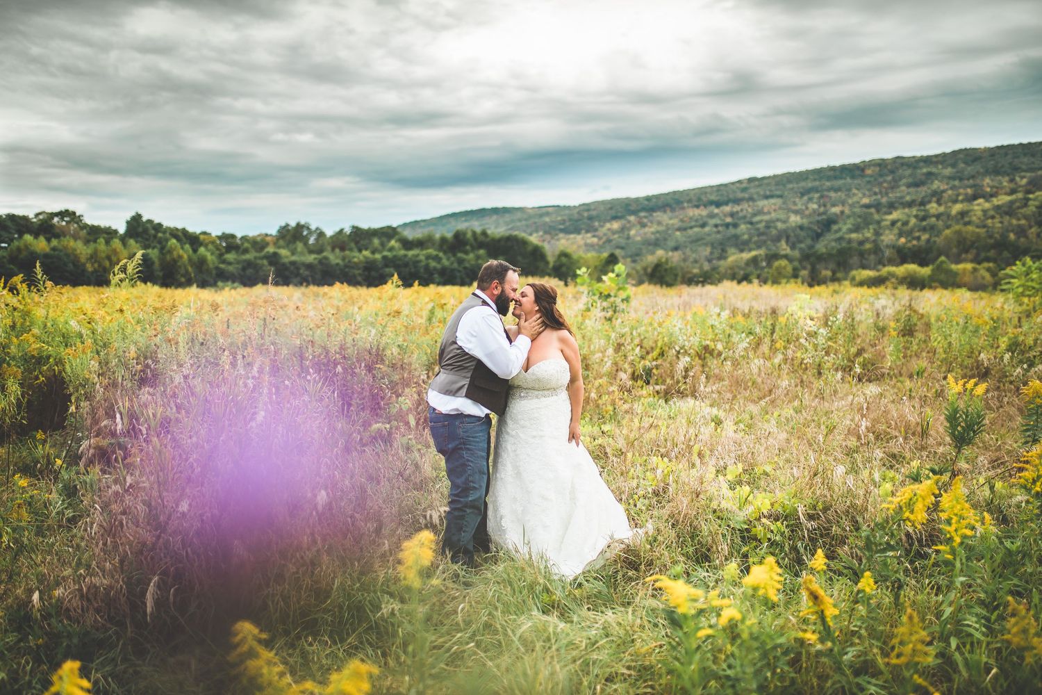 Panoramic wedding photo in a field of wildflowers under cloudy skies.
