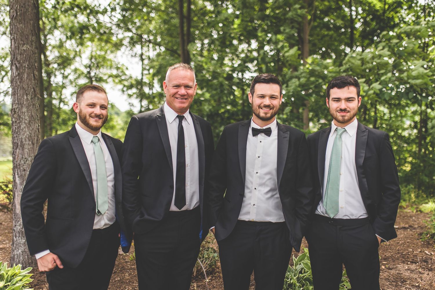 Groomsmen in black tuxedos and mint green ties pose outdoors among pine trees.