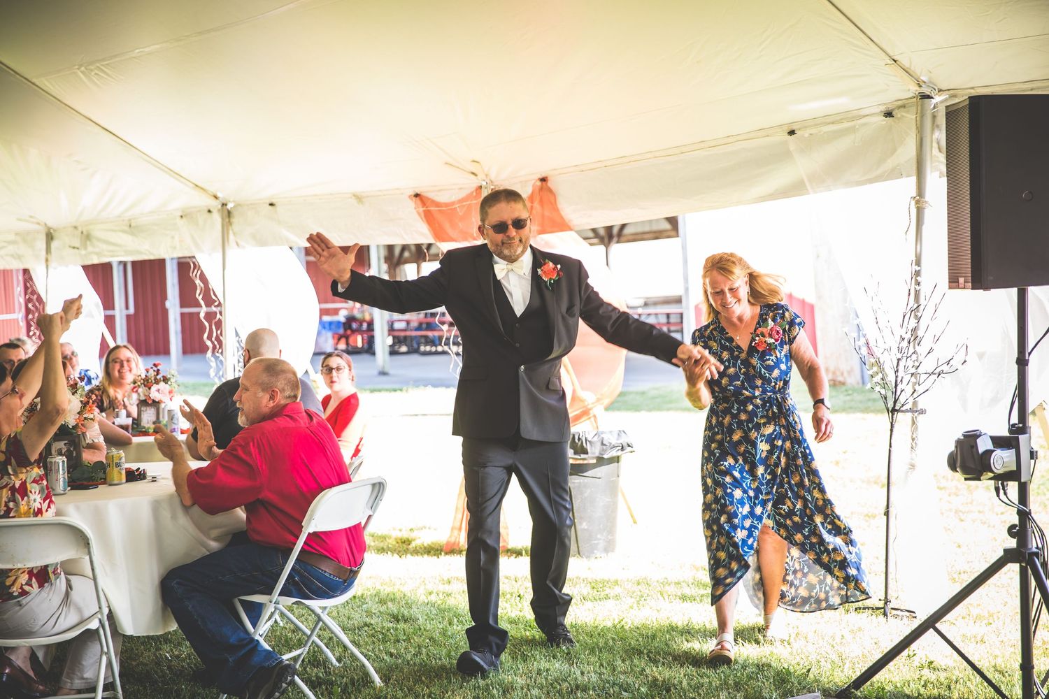 Wedding guests dance and celebrate under an outdoor tent with speakers and chairs set up for the reception.
