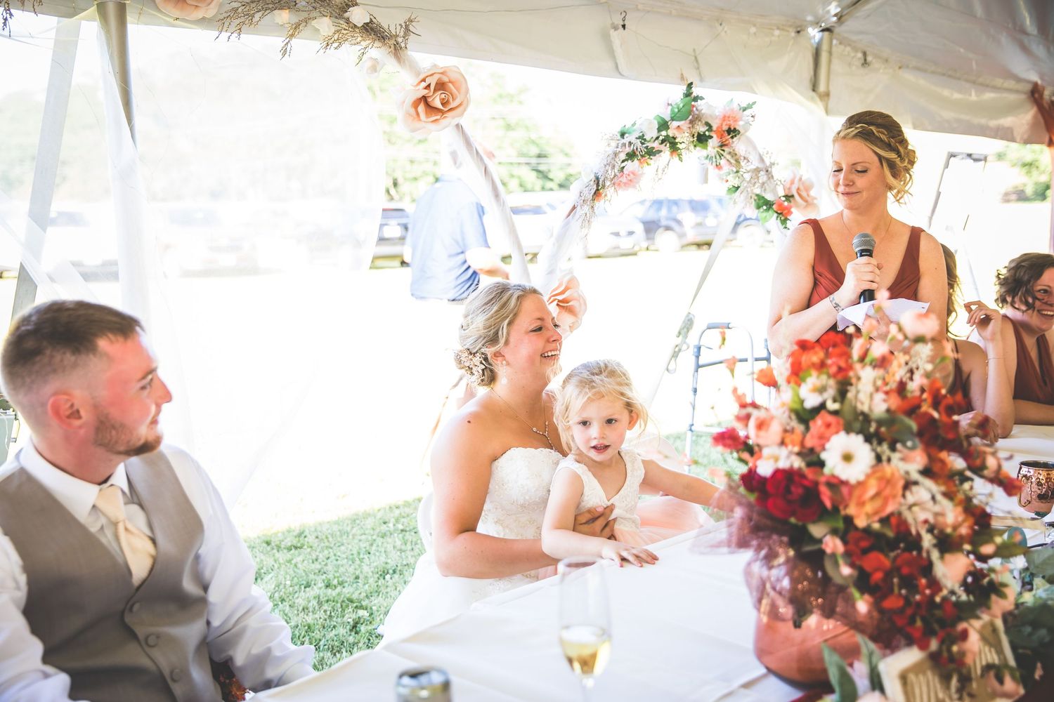 Wedding reception celebration with floral centerpieces and guests seated at tables under a tent.