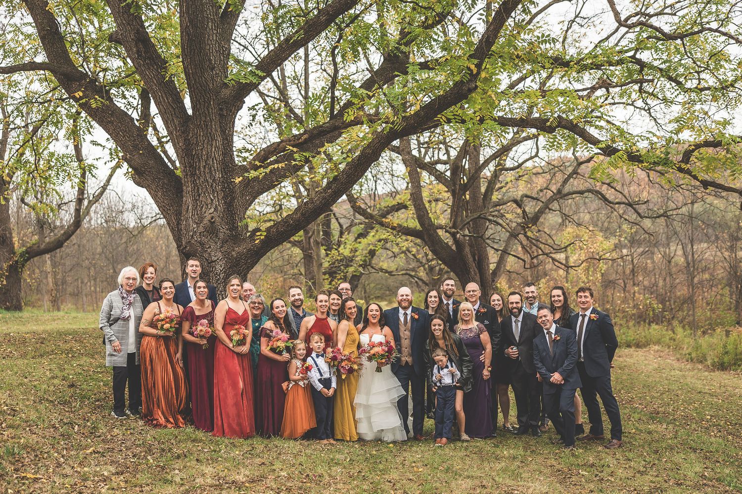 Large wedding party poses for group photo under autumn trees in outdoor setting.
