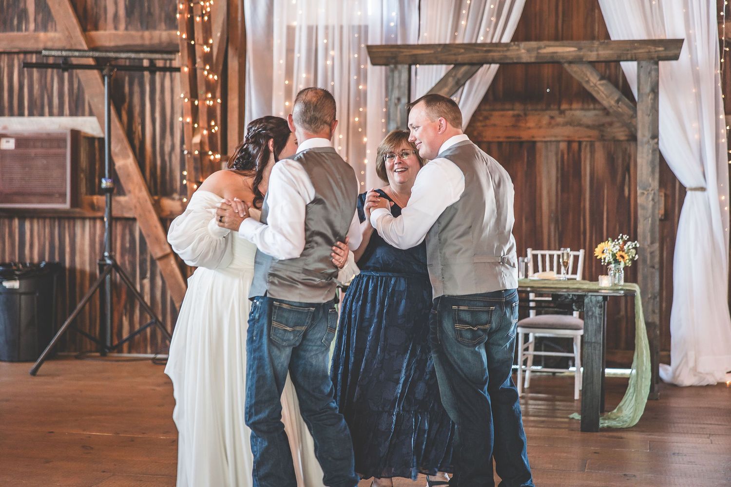 Wedding guests dance together in a warmly lit rustic barn setting with white drapes.