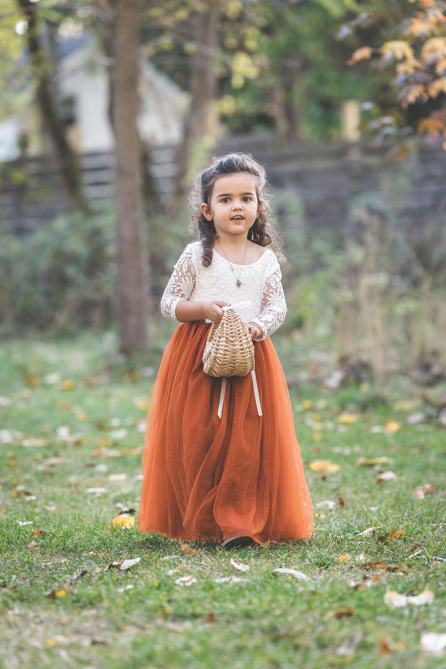 Young flower girl in white lace and orange tulle dress holds wicker basket outdoors.