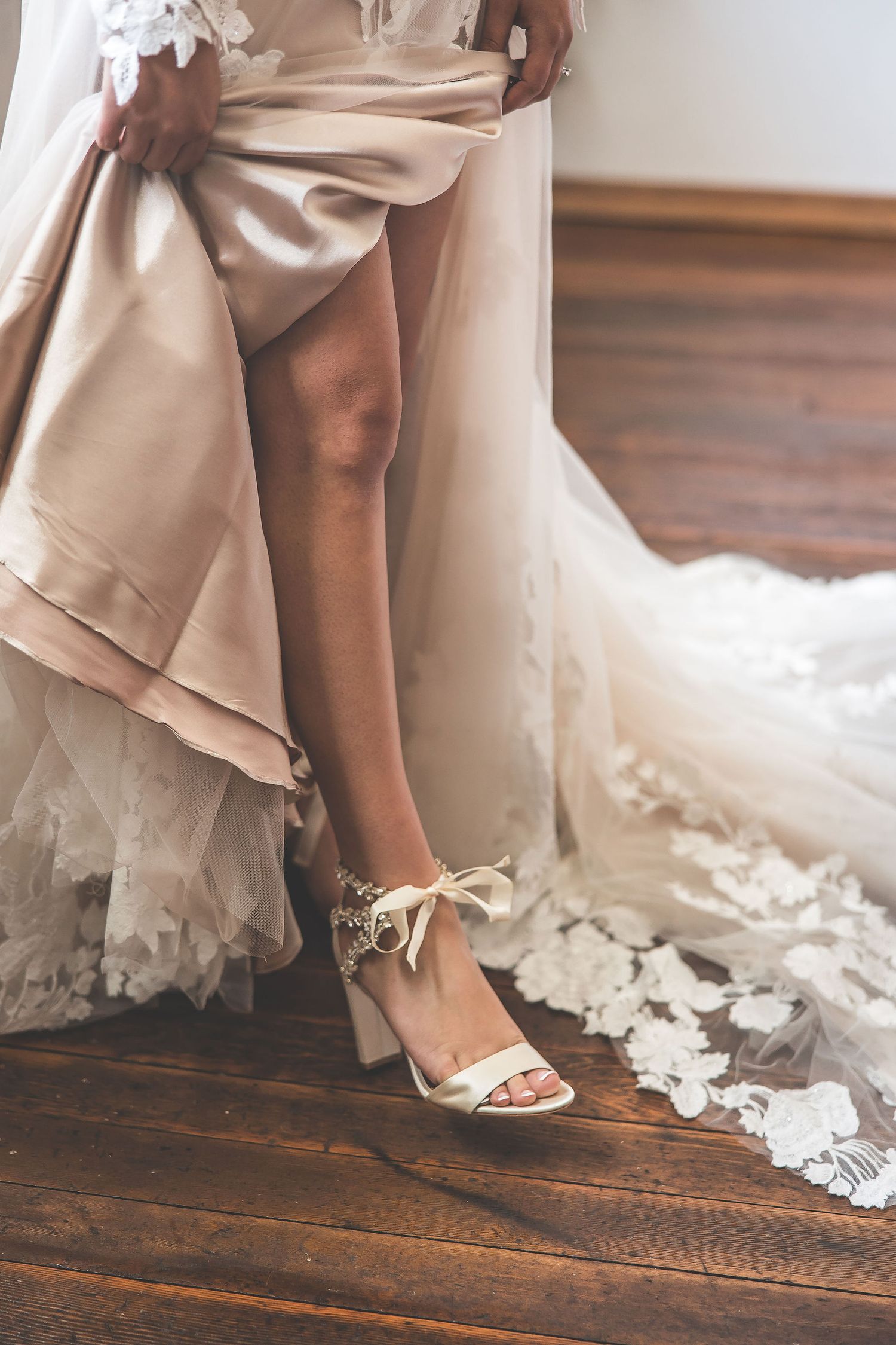 Elegant bridal shoes peek out beneath layers of delicate lace wedding dress on hardwood floors.