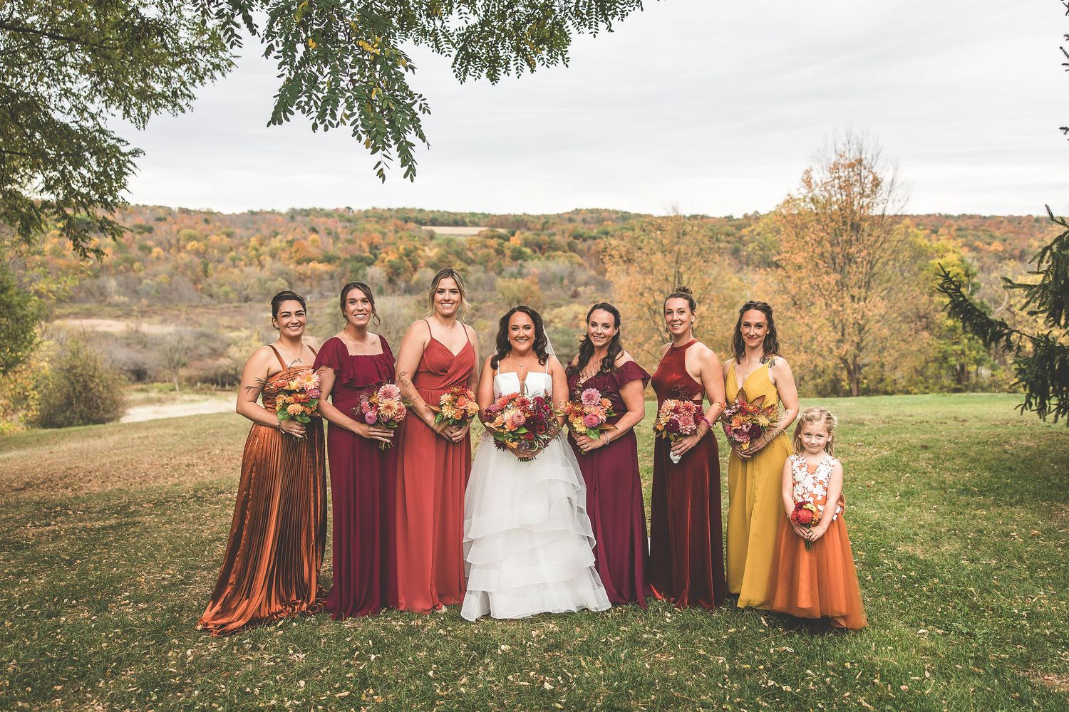 Wedding party in burgundy and orange dresses poses together in an autumn field with rolling hills.