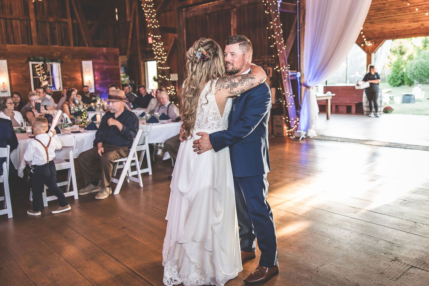 Wedding party dancing and celebrating in rustic barn decorated with white drapes and ambient lighting.