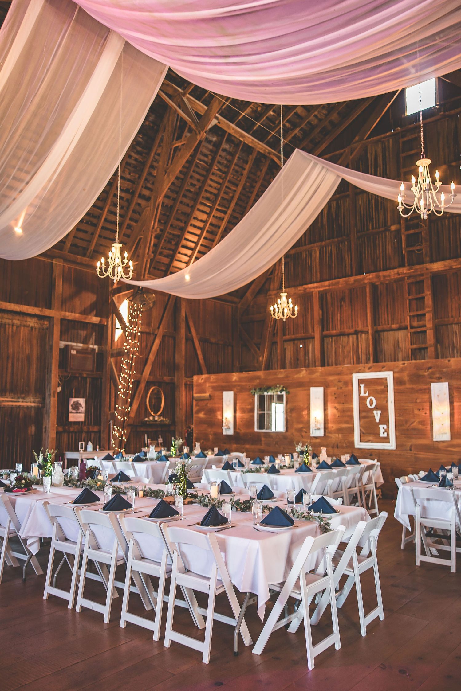 White folding chairs arranged in rows inside a barn with elegant fabric draping and chandeliers.