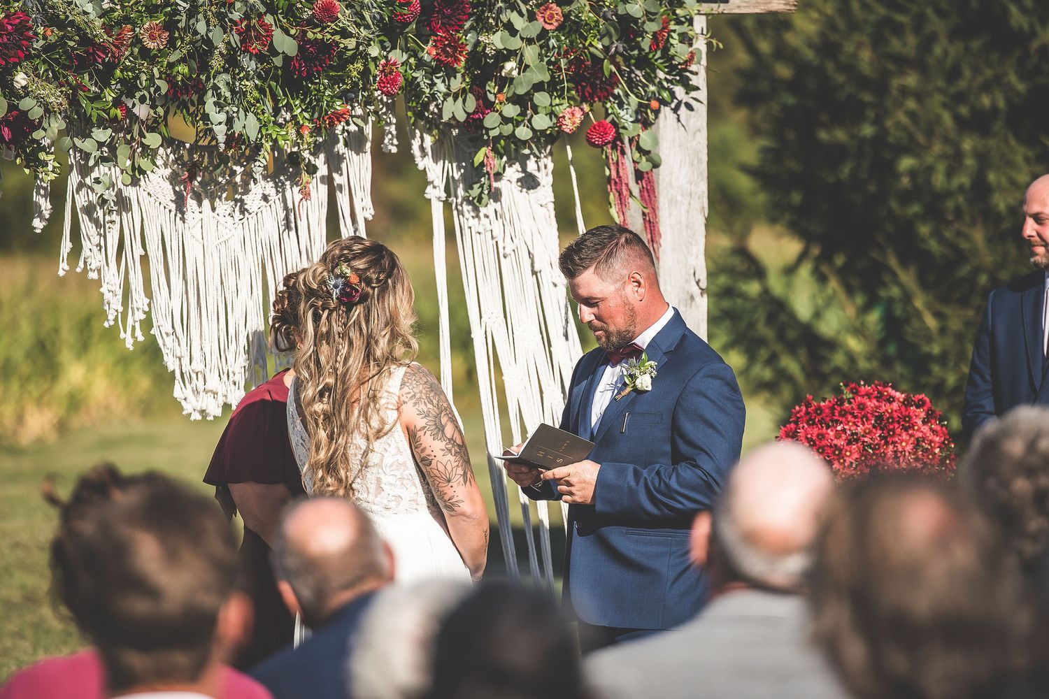 Wedding ceremony with floral decorations and white macrame backdrop in an outdoor setting.