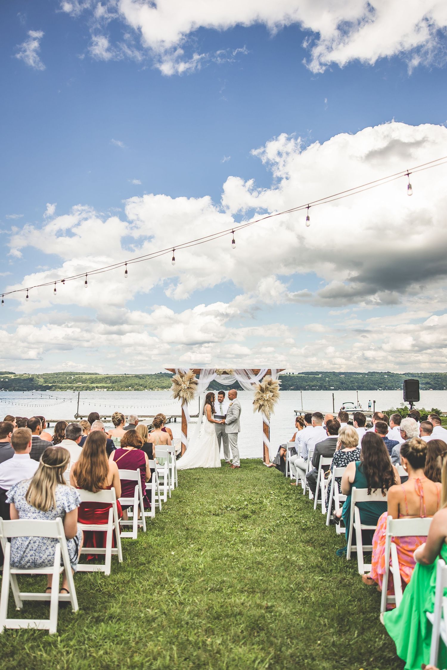 White chairs set up on grass lawn for waterfront wedding ceremony under blue skies with string lights.