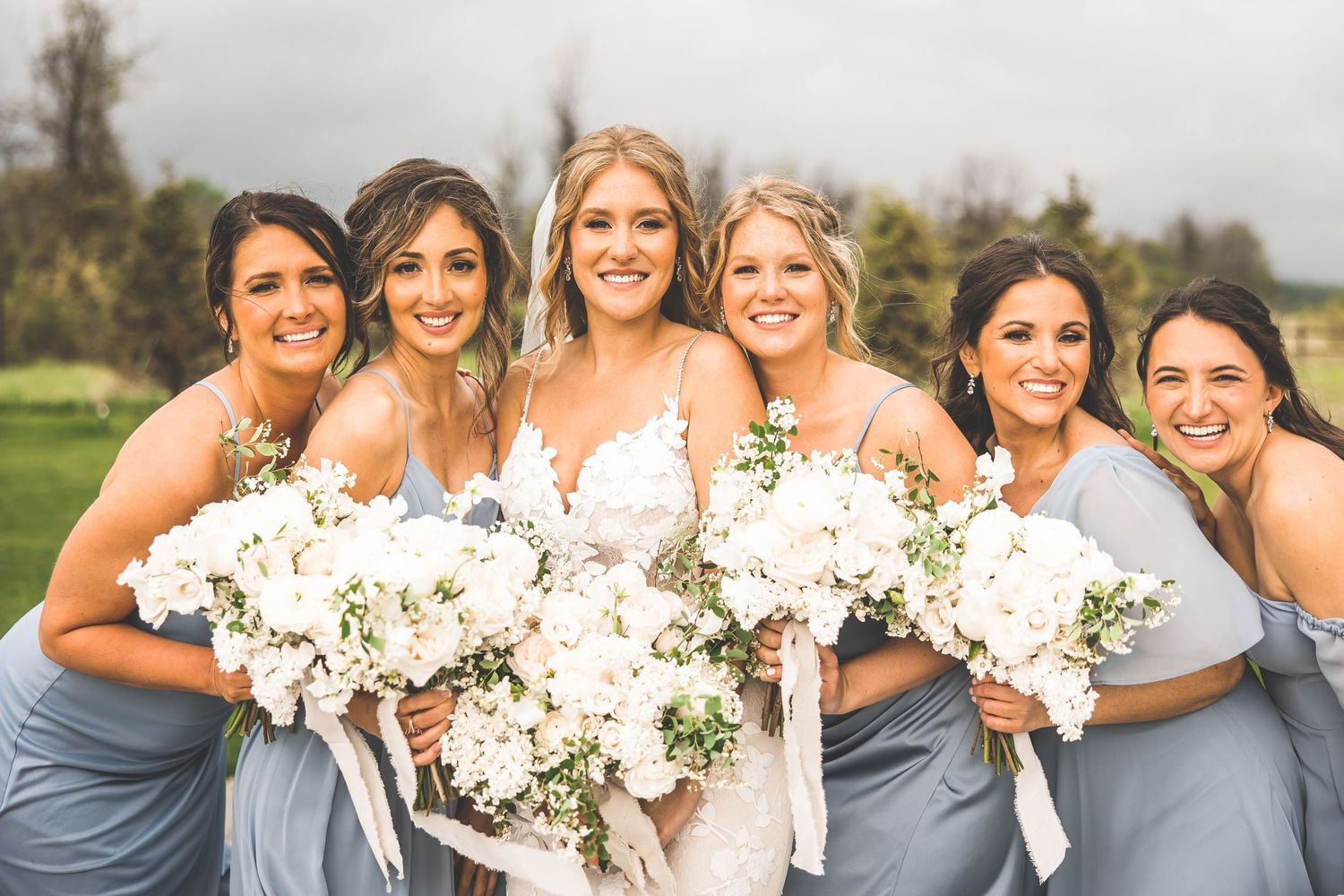 Bridesmaids in dusty blue dresses hold elegant white floral bouquets while smiling outdoors.