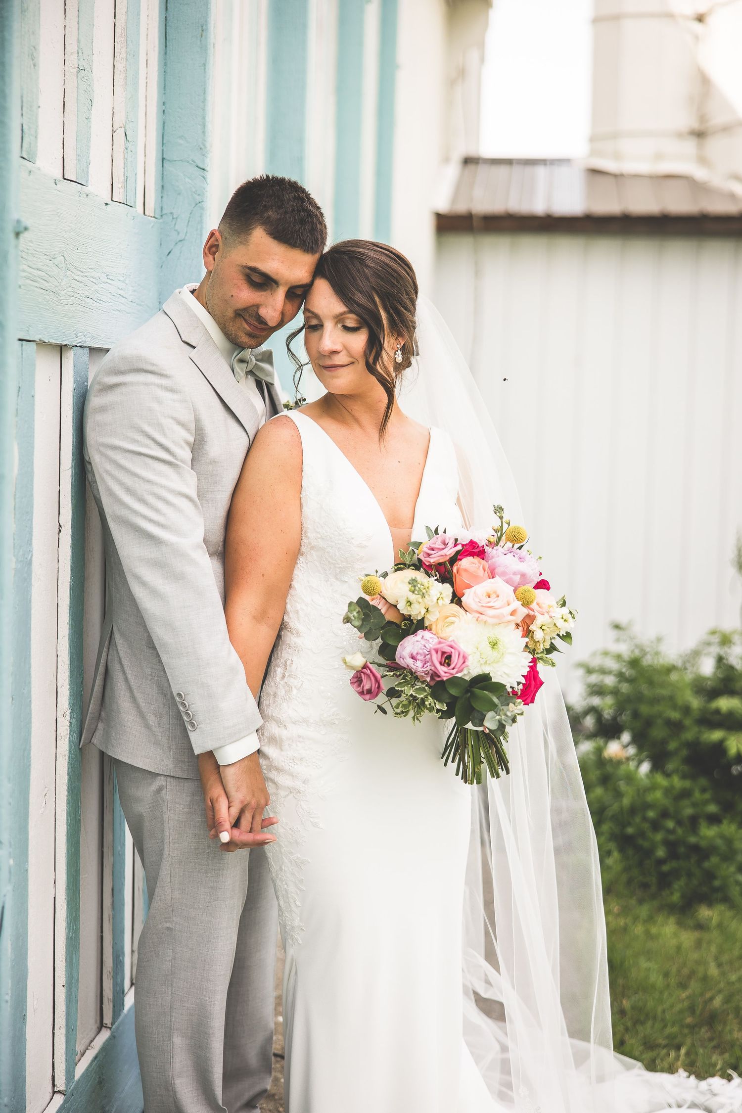 A romantic wedding couple poses in front of a light blue wall while holding a pink and white floral bouquet.