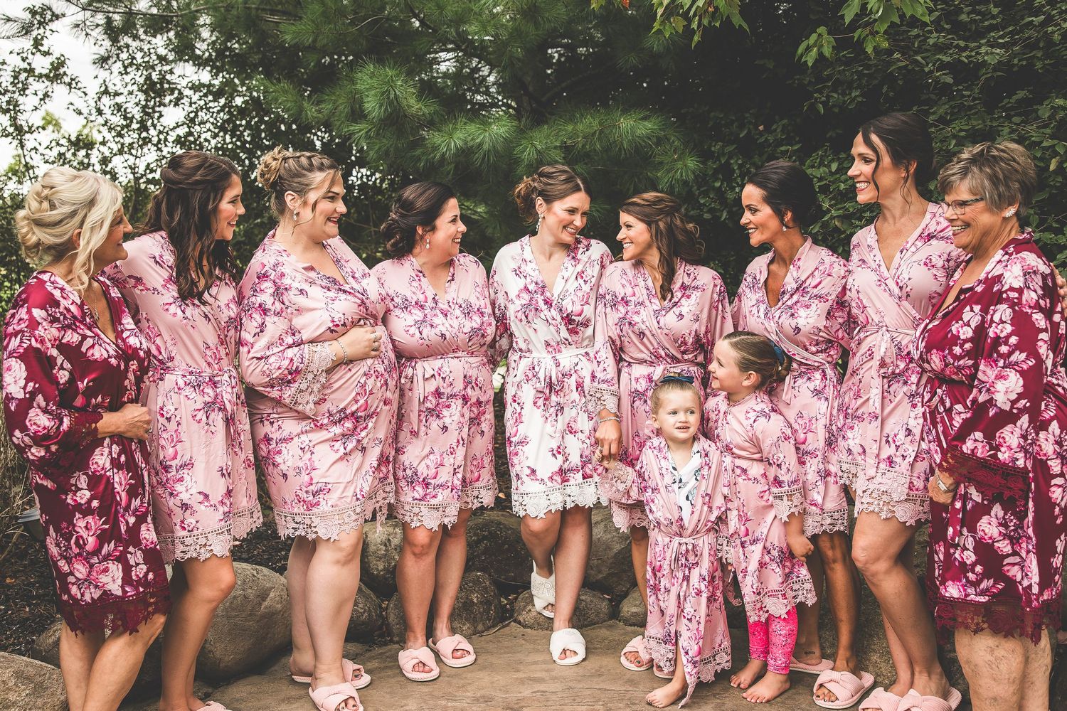 Bridal party in matching pink floral robes laughing together outdoors.