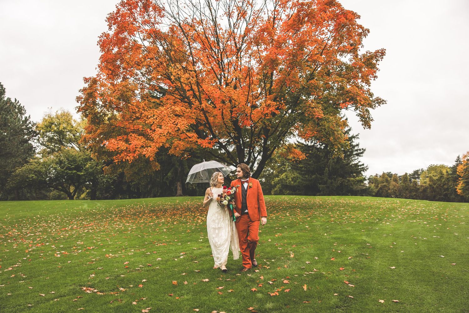 Couple with umbrella stands beneath stunning orange maple tree on wedding day.