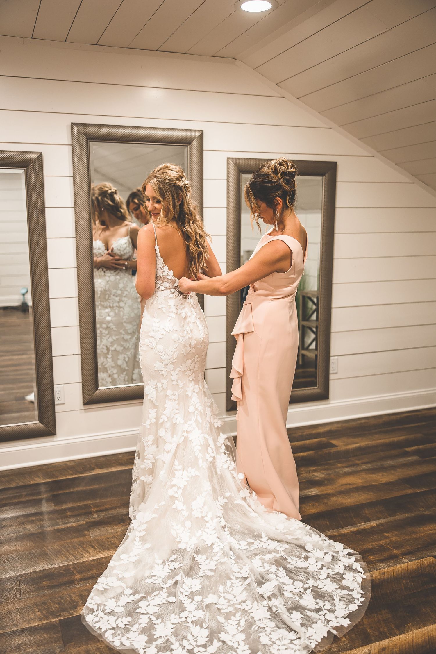 Bridesmaid in pink dress helping bride with fitted lace wedding gown.