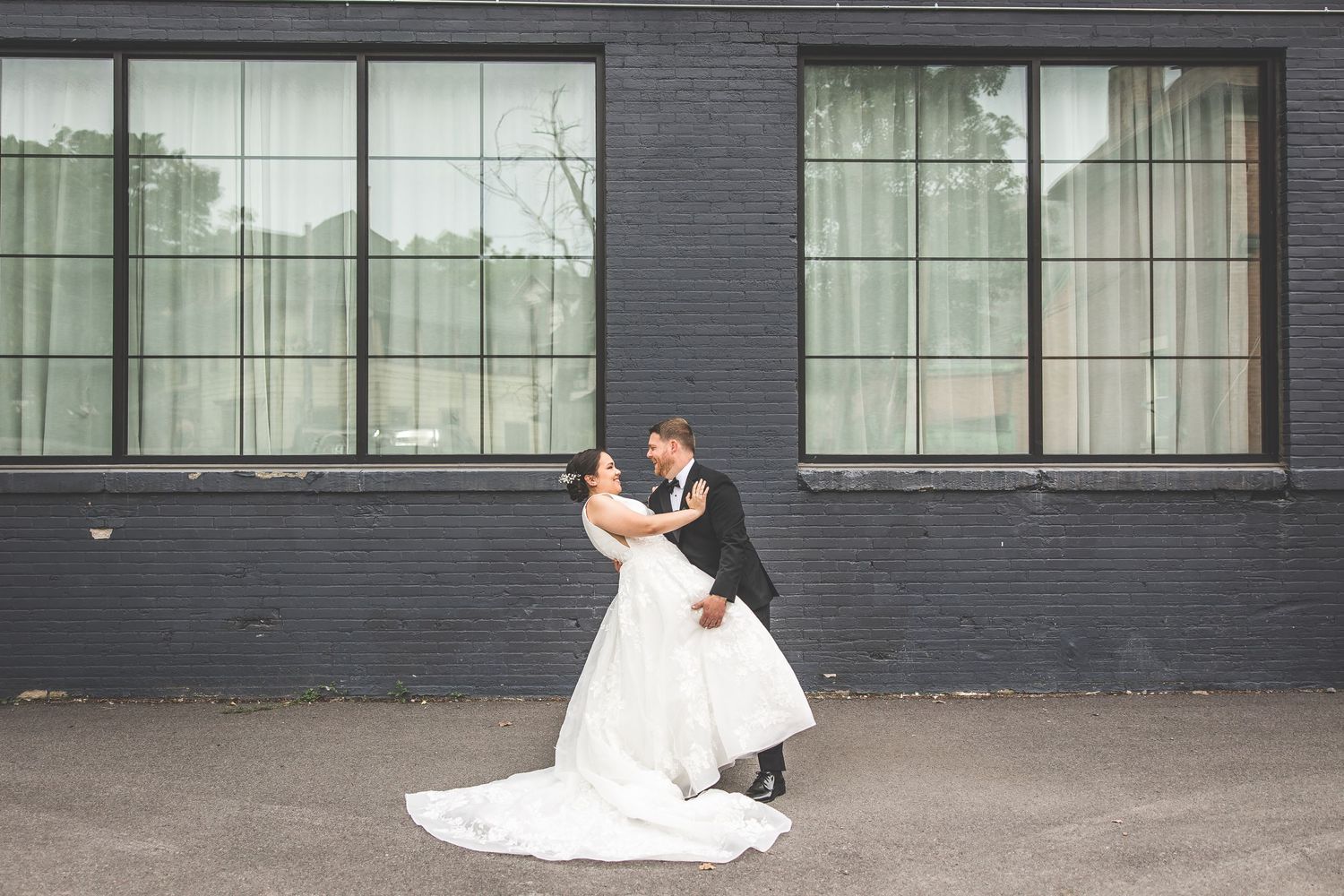 Wedding couple embraces while standing beside a dark painted building exterior.