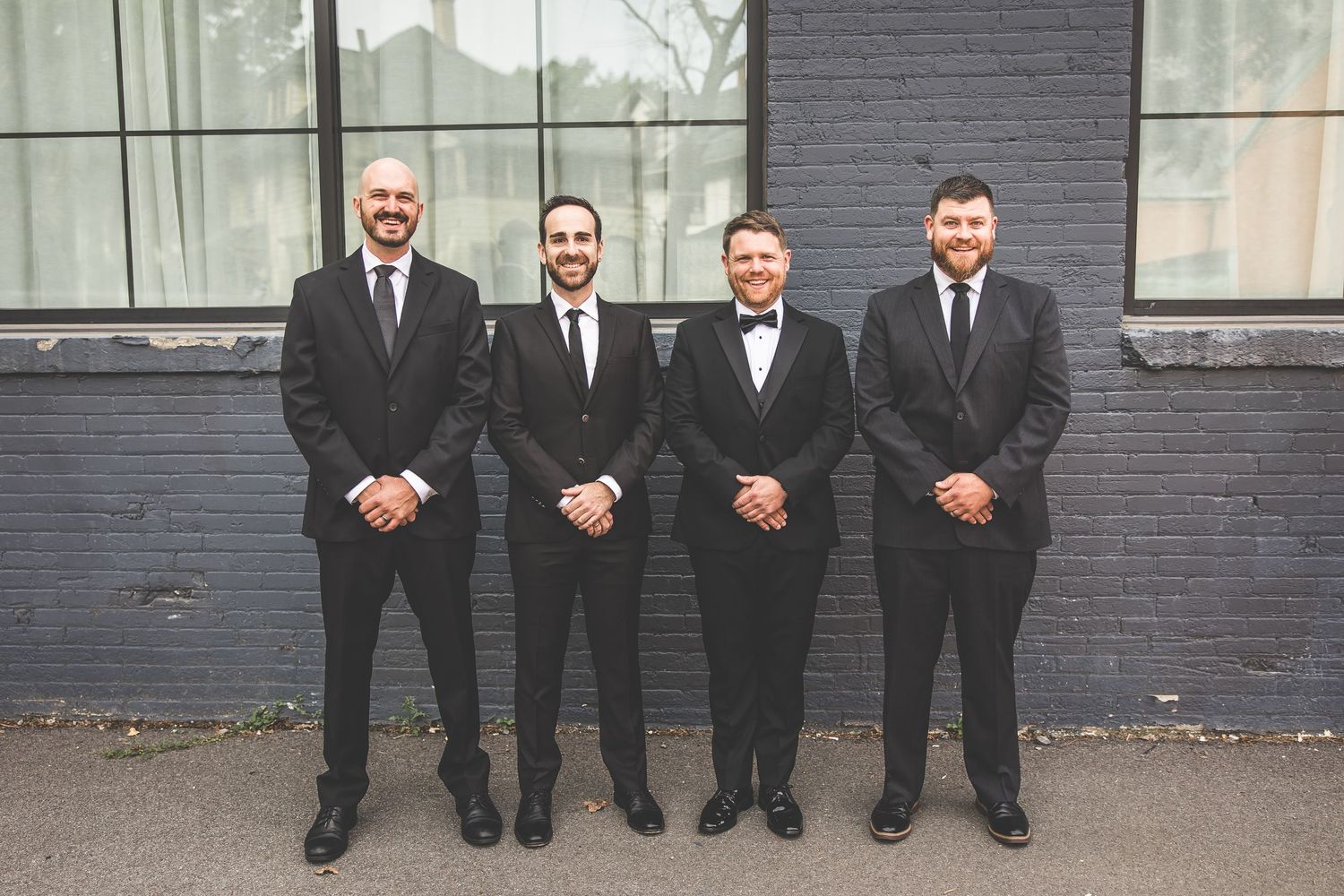 Groomsmen in black suits pose casually against a dark brick wall outdoors.