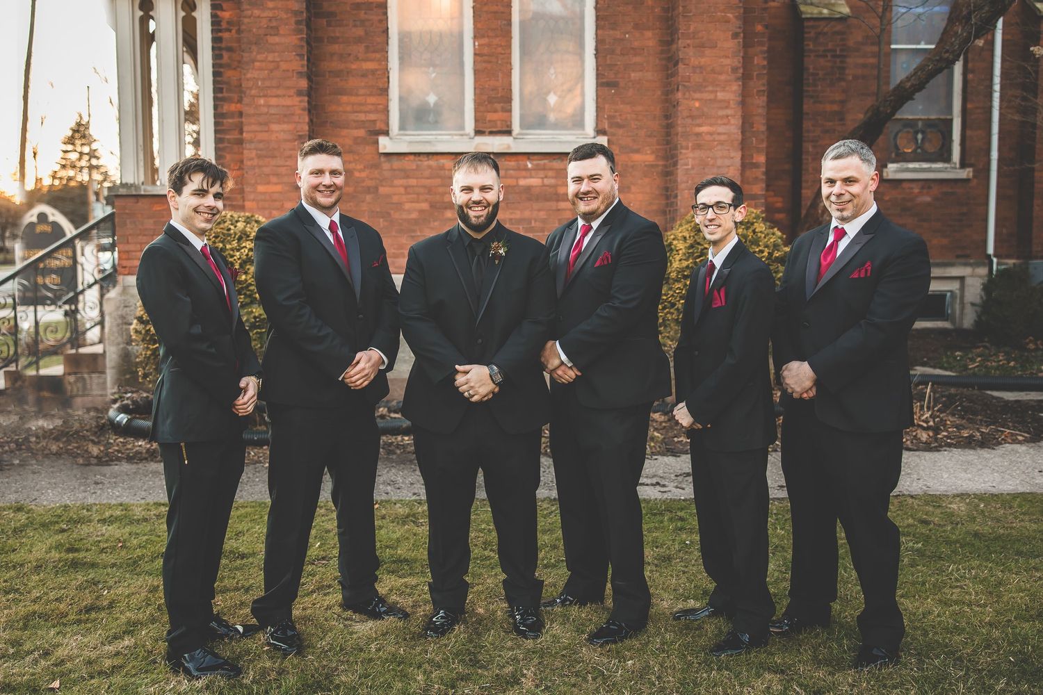 Wedding party members lined up in formal attire against brick building backdrop.