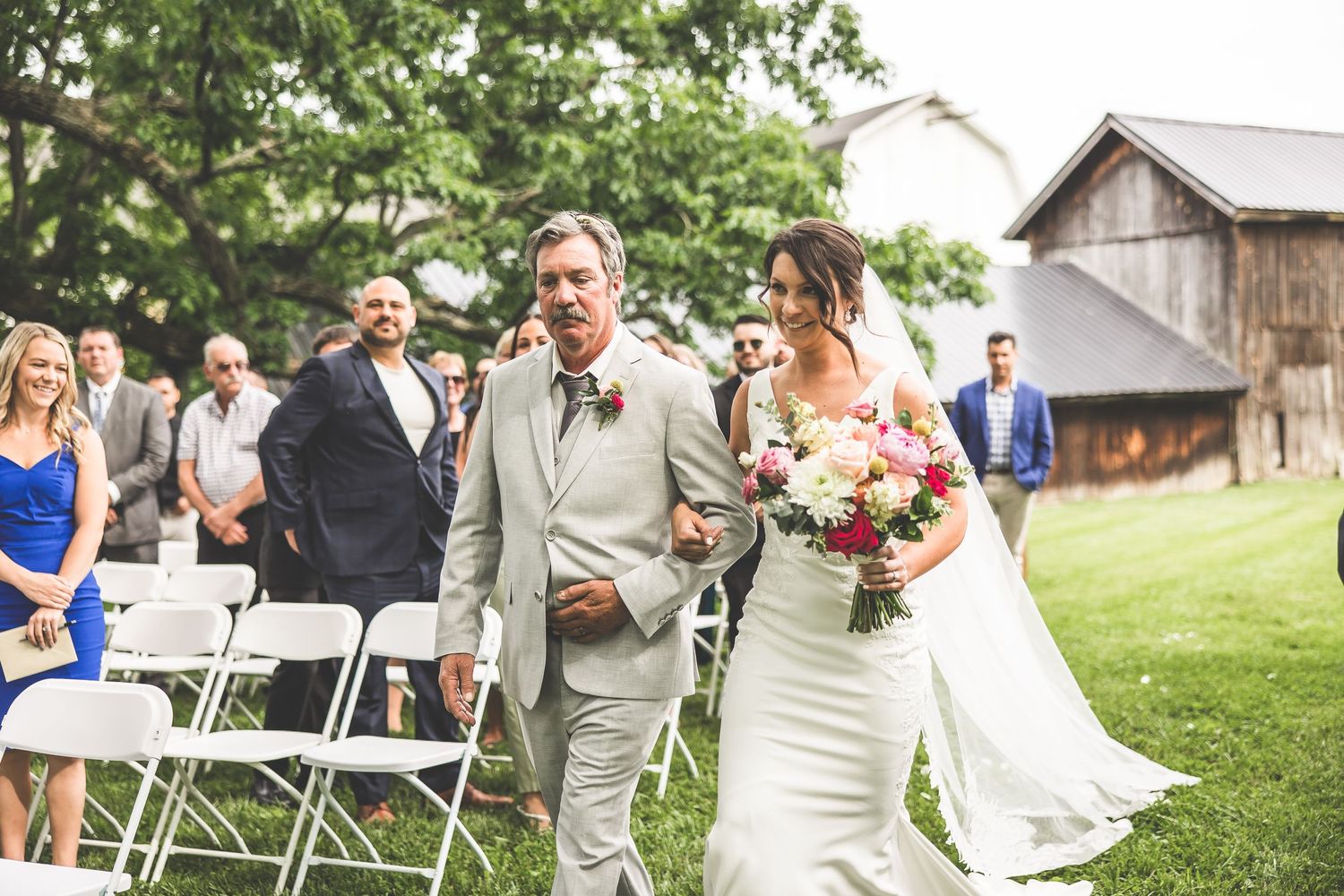 Wedding ceremony processional on grassy lawn with rustic barn in background.