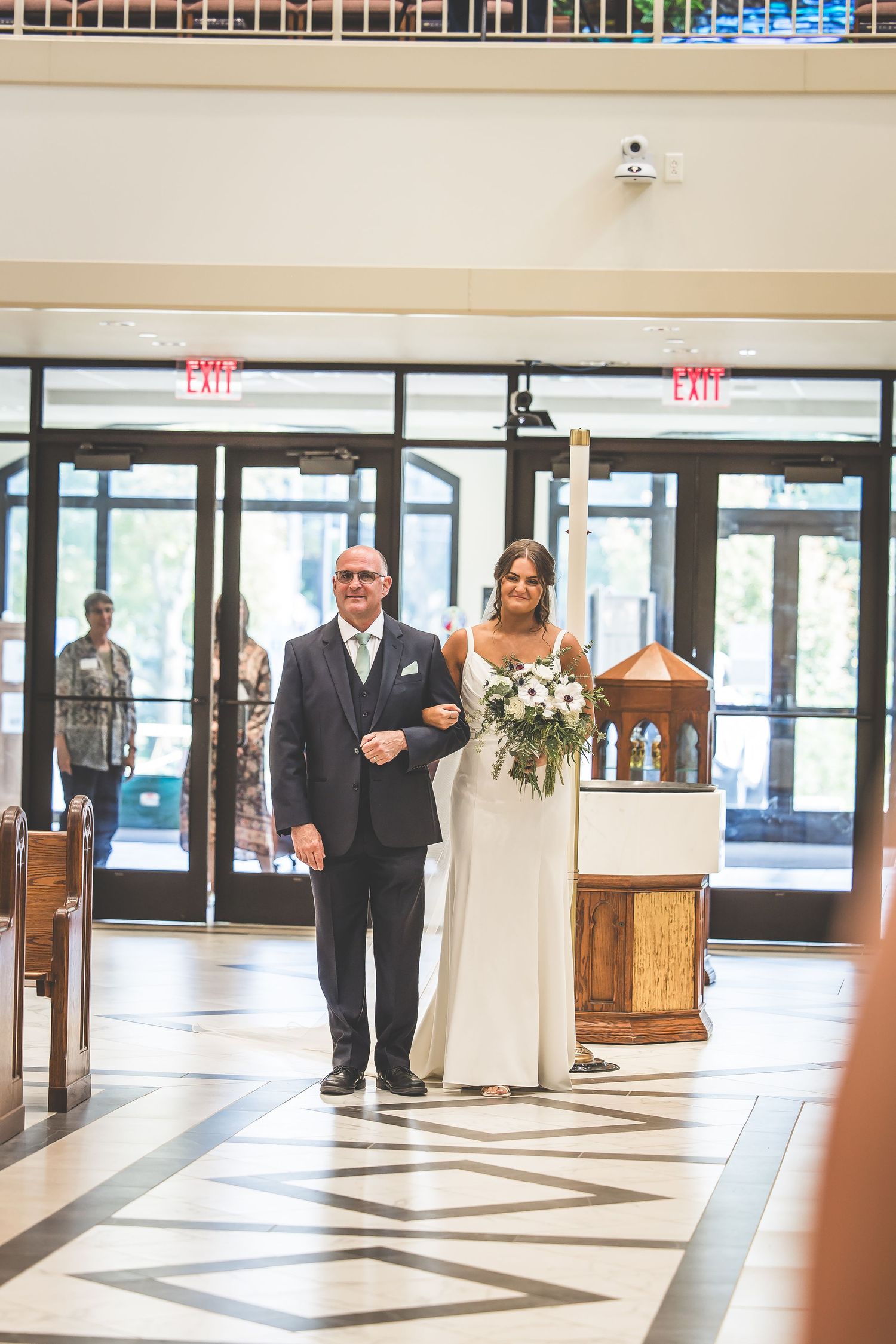 Wedding party members make their entrance through glass doors into a modern church foyer.