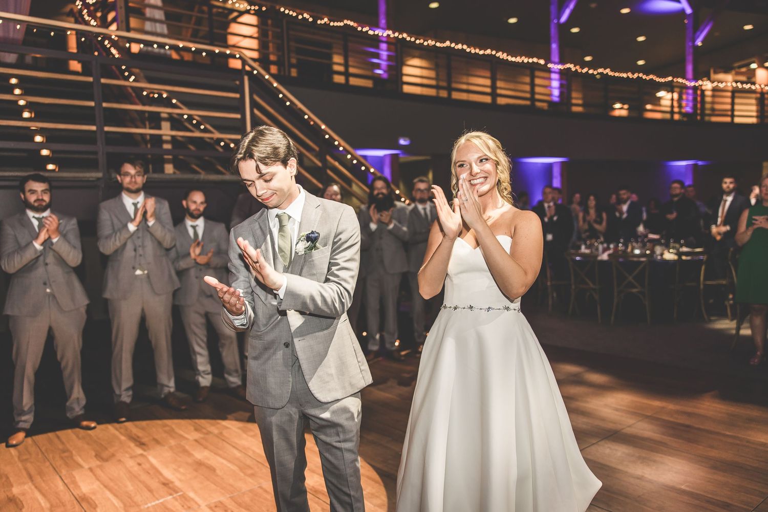 Wedding celebration on wooden dance floor with wedding party in grey suits applauding.