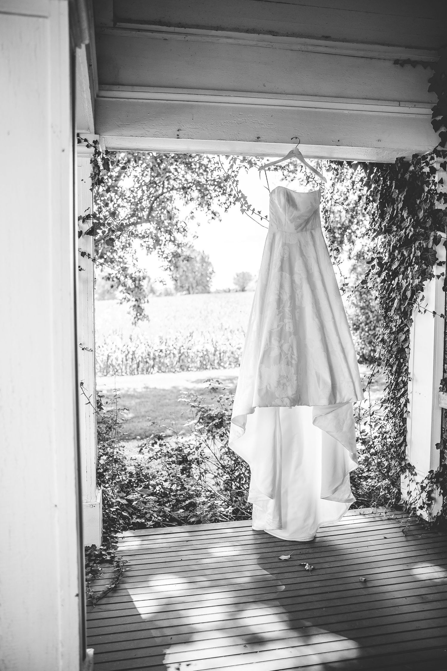 Elegant white wedding dress hanging in doorway with natural light streaming through surrounding foliage.