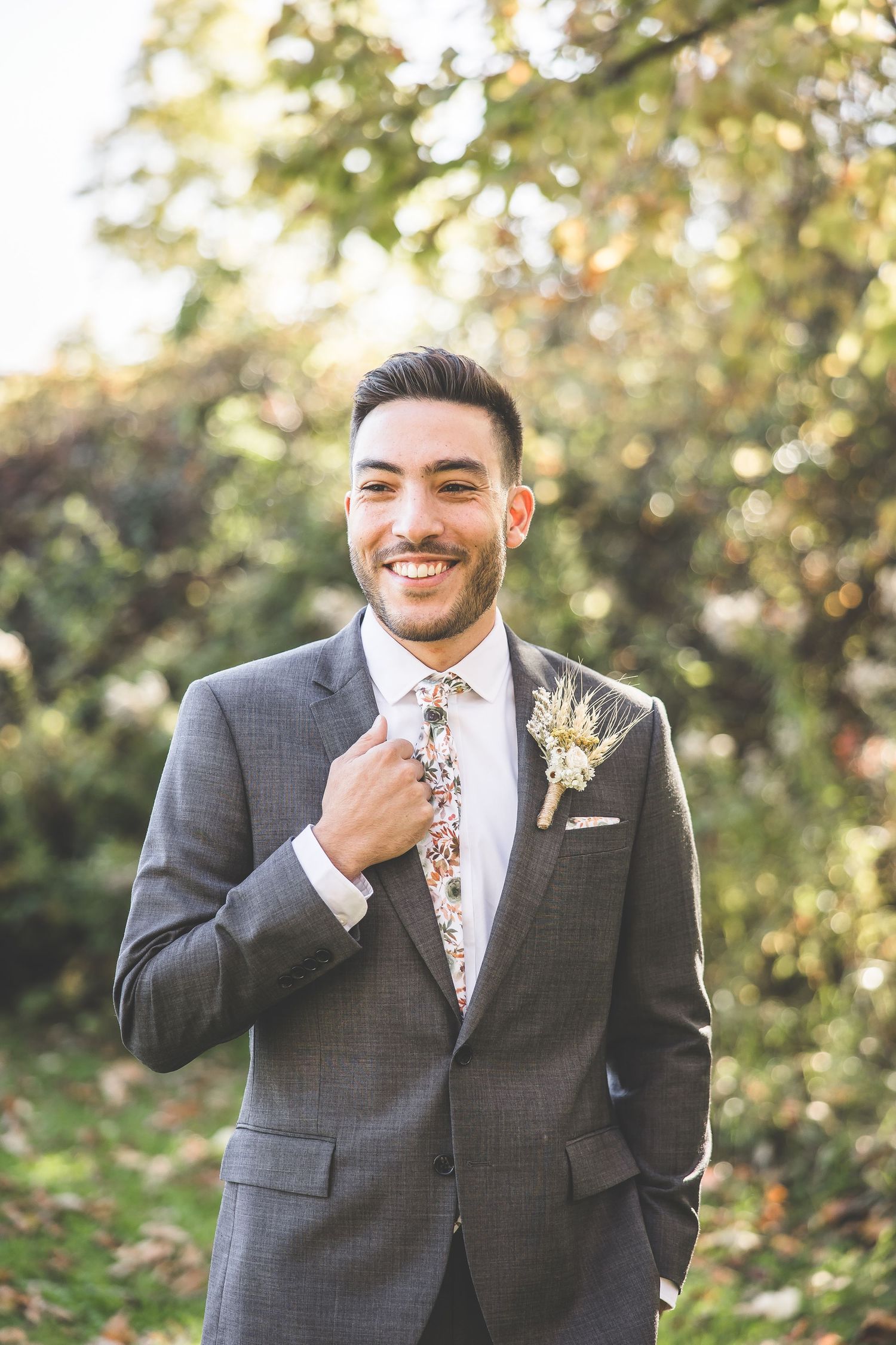 Groom in gray suit with floral tie and boutonniere smiling outdoors.
