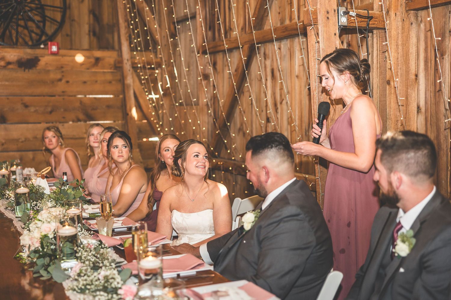 Wedding reception dinner setup in rustic barn with wooden tables and floral centerpieces.