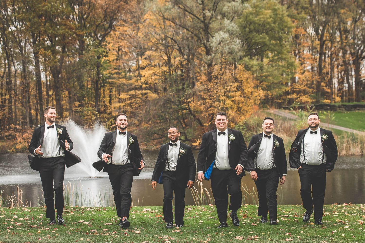 Wedding party poses playfully in formal attire against autumn foliage backdrop.