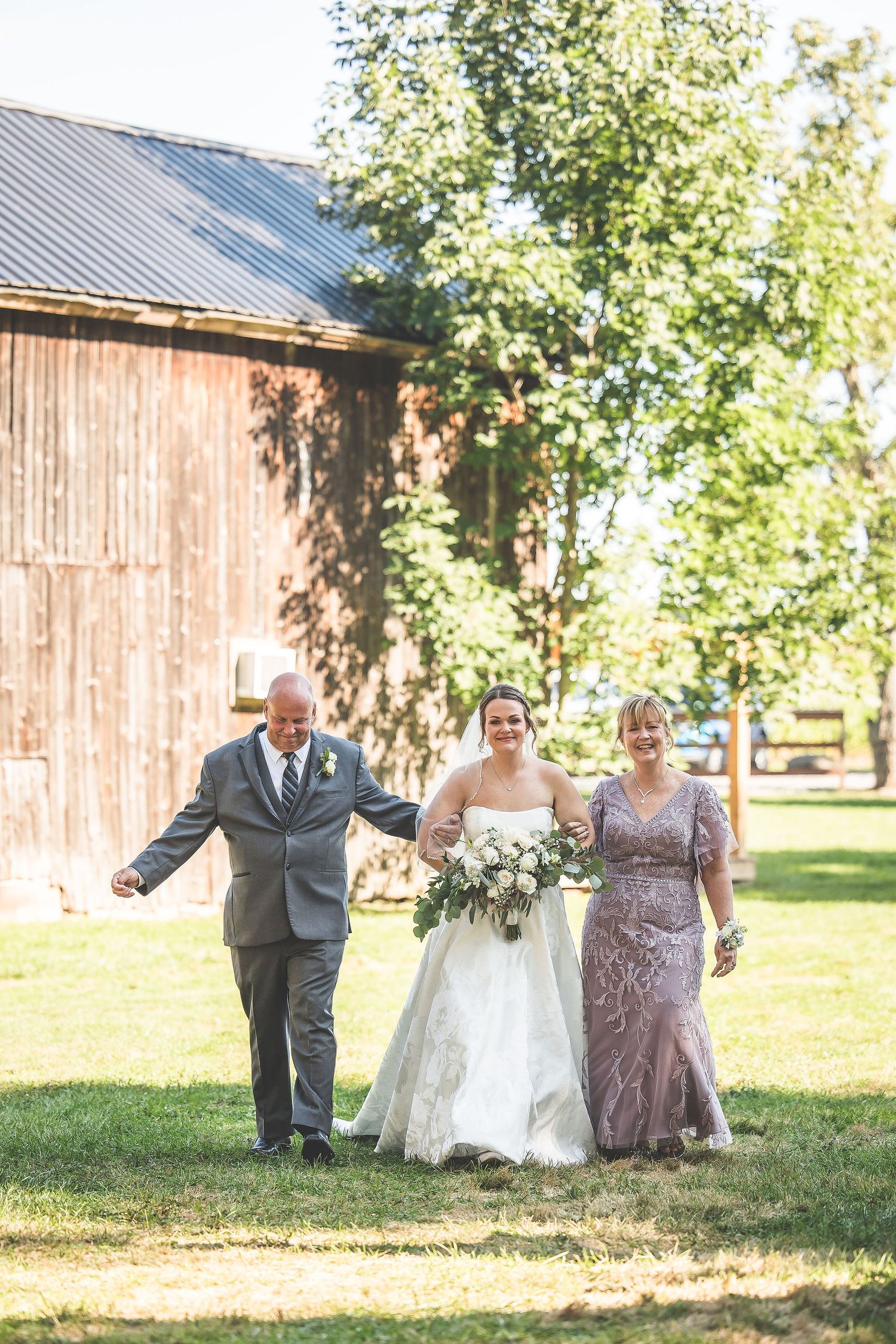 Outdoor wedding ceremony with rustic barn backdrop and sunlit trees in a natural setting.