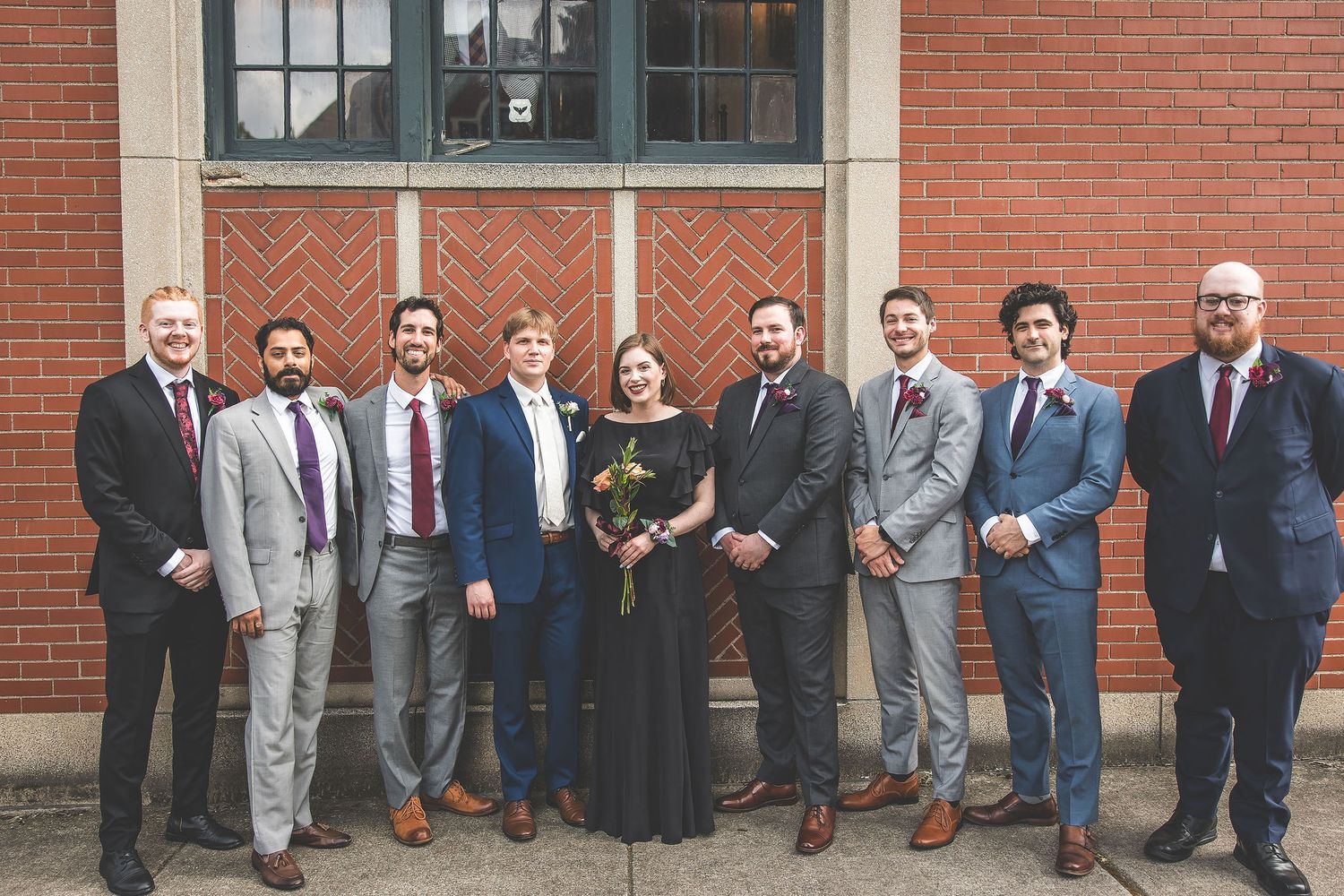 Wedding party members in suits and formal attire posed along brick building exterior.