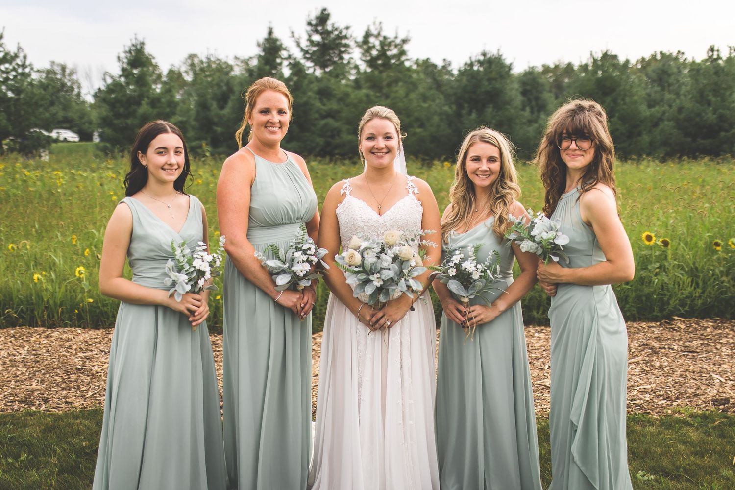 Bridesmaids in sage green dresses stand together holding bouquets in an outdoor setting.