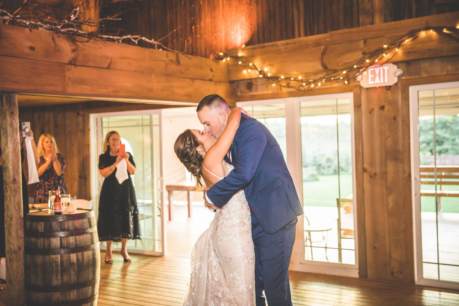 A romantic first kiss between newlyweds in a rustic barn venue with string lights and wooden barrels.