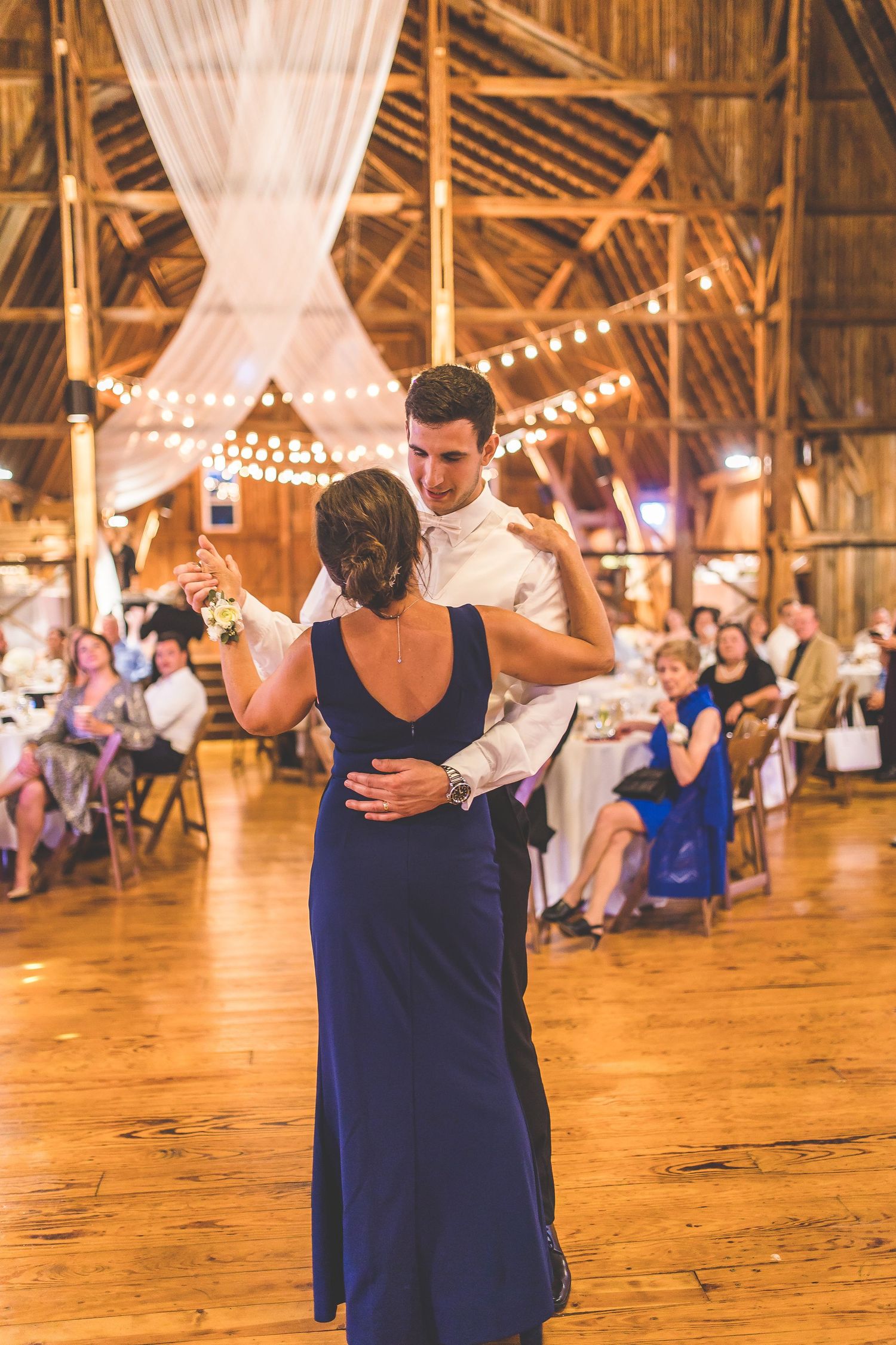 Couples sharing dances on a wooden floor in a barn decorated with string lights and white draping.