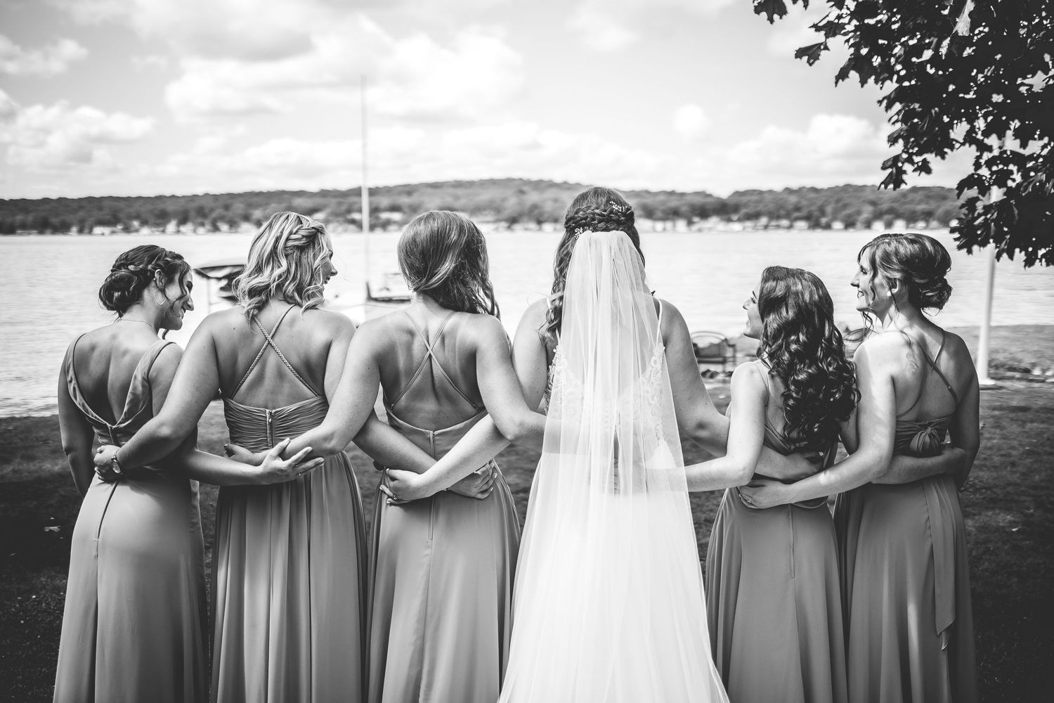 Bridesmaids in sage green dresses stand arm-in-arm overlooking a lake.