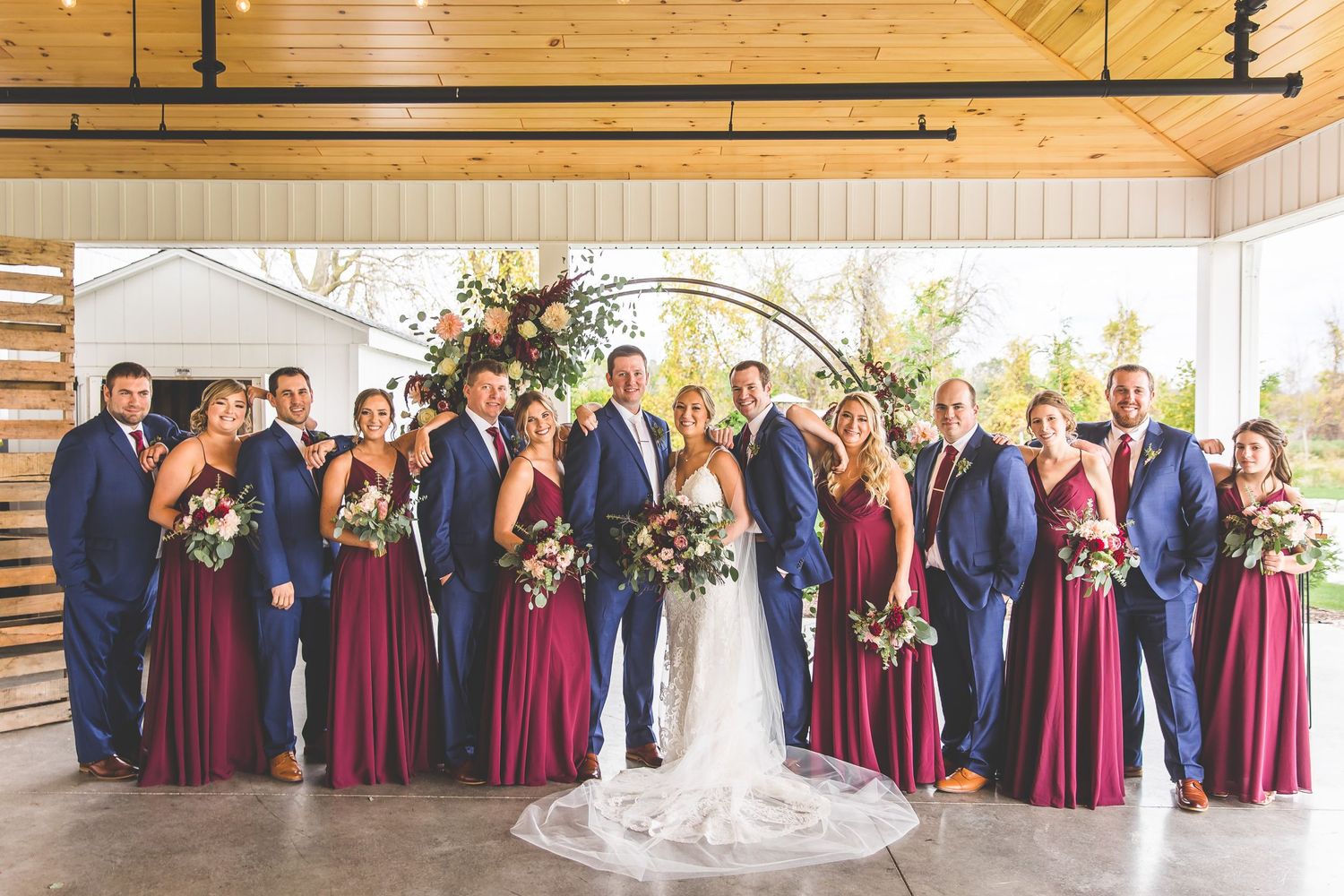 Wedding party photo with burgundy and navy color scheme under modern venue arch.