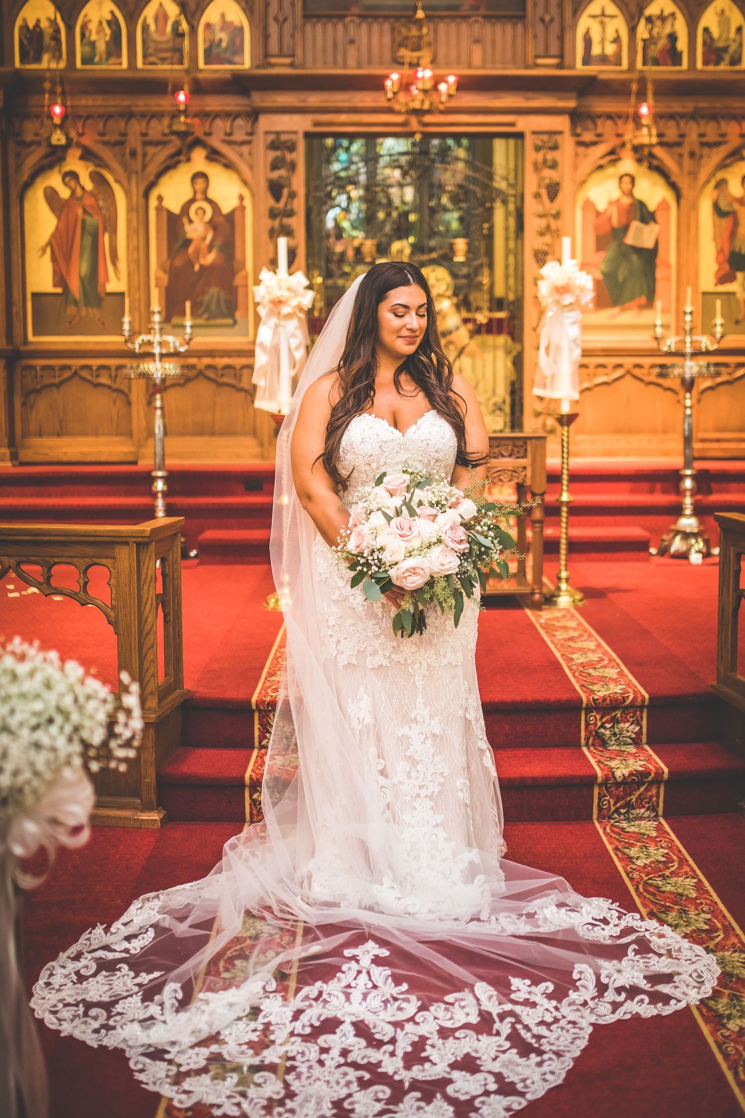 A bride in a lace wedding dress with long train stands in an ornate church with religious icons and candles.