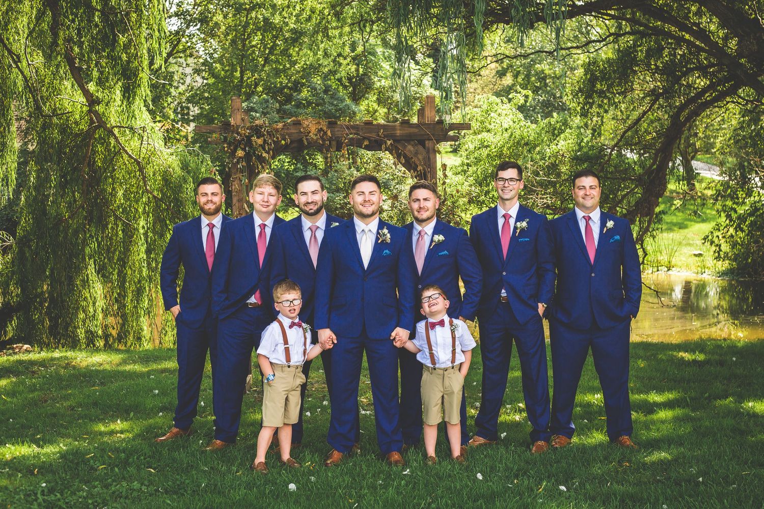 Groomsmen and ring bearers in matching navy suits pose under willow trees.