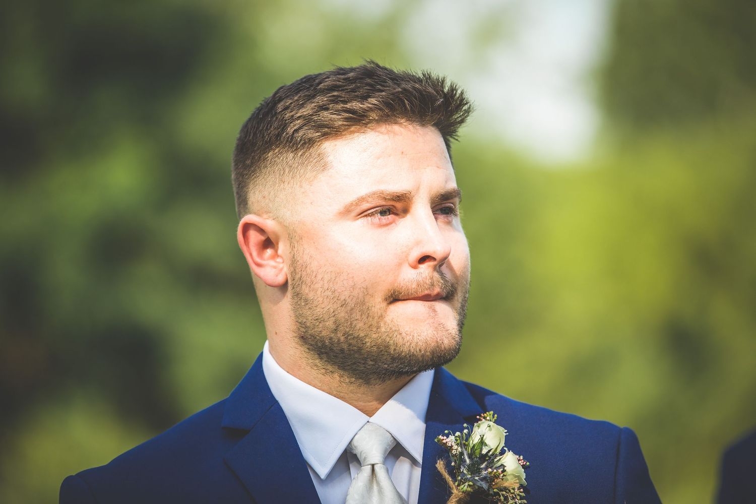 Groom in navy suit with boutonniere looking emotional outdoors.