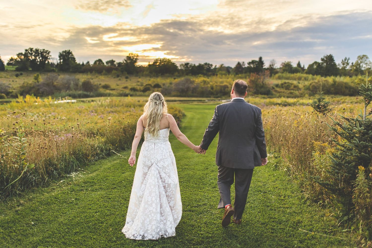 Newlyweds stroll together through a rustic field with wildflowers at dusk.