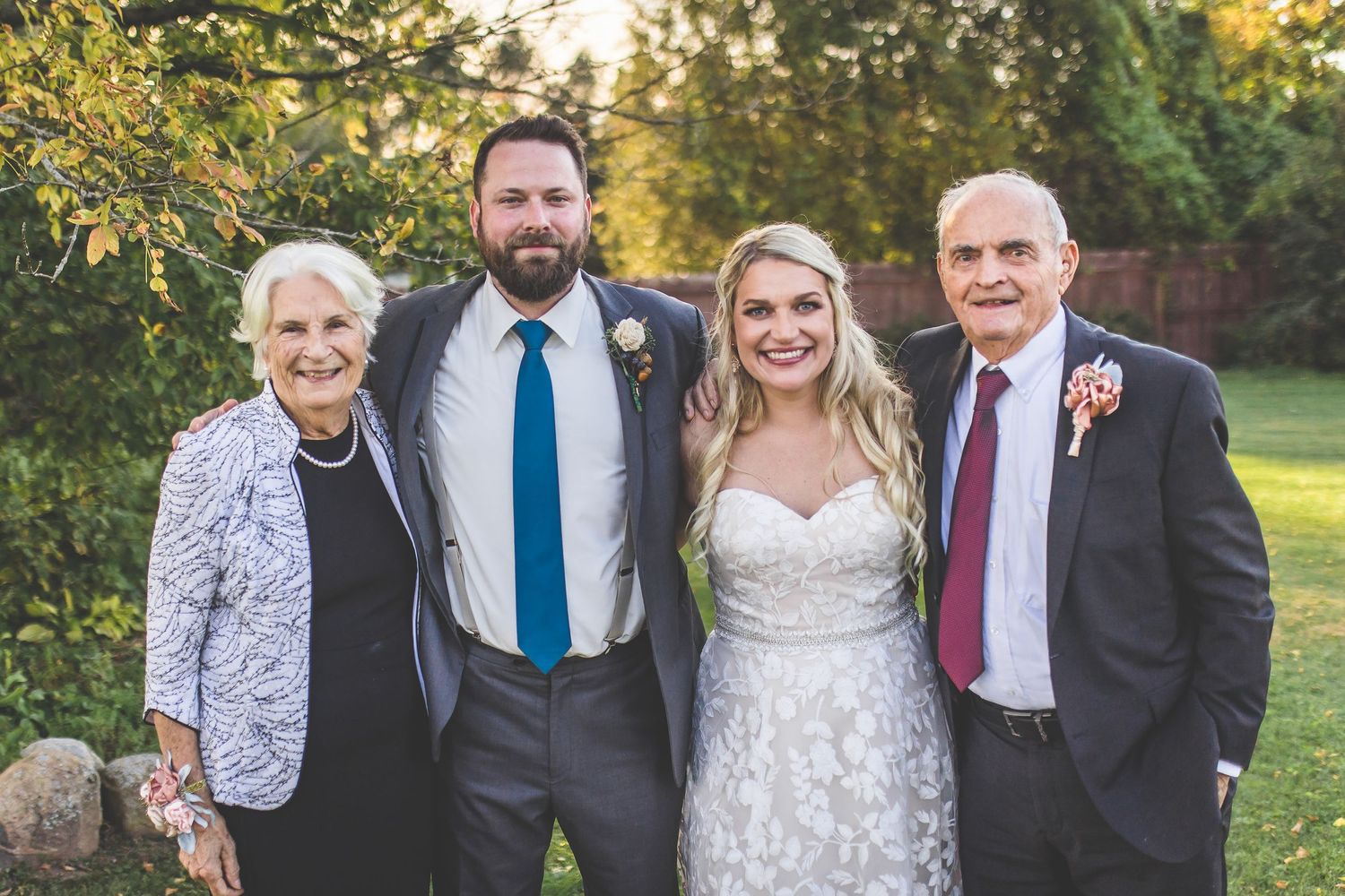 Wedding party poses outdoors in formal attire against natural greenery at sunset.