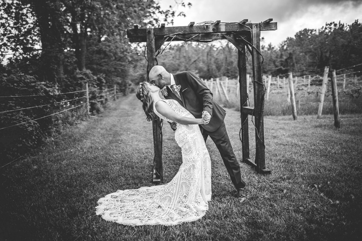 A dramatic black and white wedding photo showing a romantic dip under a rustic wooden arch.