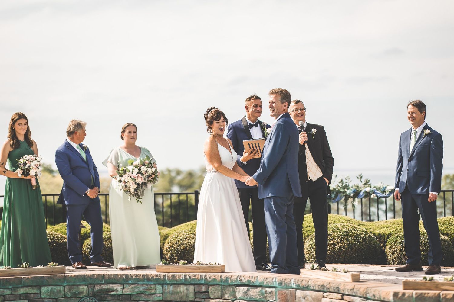 Wedding party gathered outdoors on stone patio with swimming pool and scenic landscape in background.
